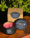 A Feminist Rage soy wax candle in a matte black tin with pink wax, shown alongside its kraft paper box and lid on a wooden surface with green foliage in the background.