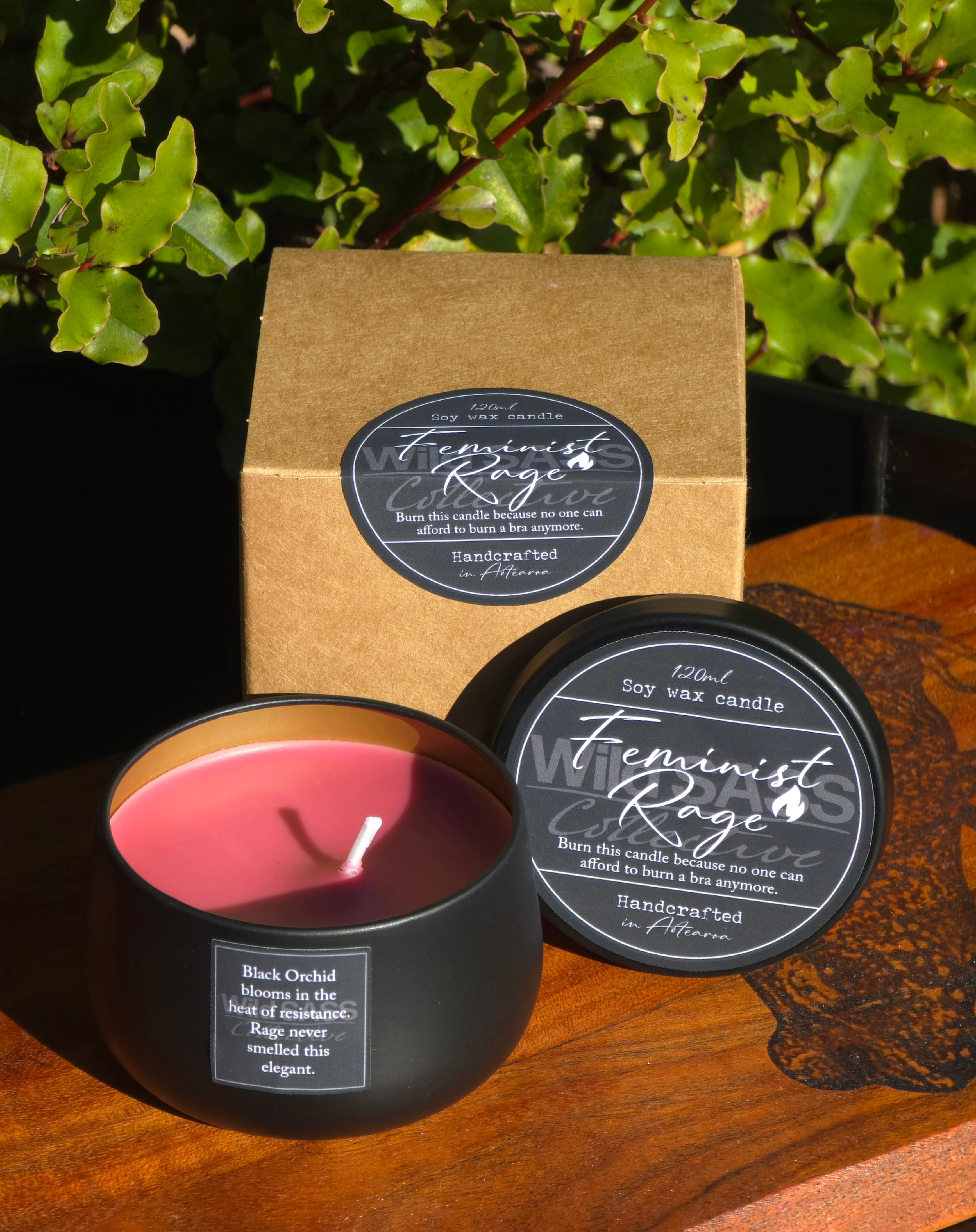 A Feminist Rage soy wax candle in a matte black tin with pink wax, shown alongside its kraft paper box and lid on a wooden surface with green foliage in the background.